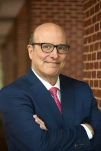 A headshot of a man in a blue suit, standing with his arms crossed against a blurry brick background.